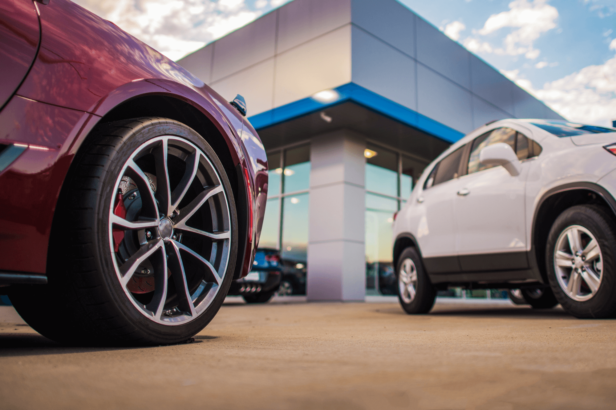 Cars parked outside a modern car dealership, including a red vehicle in the foreground and a white SUV near the showroom entrance.
