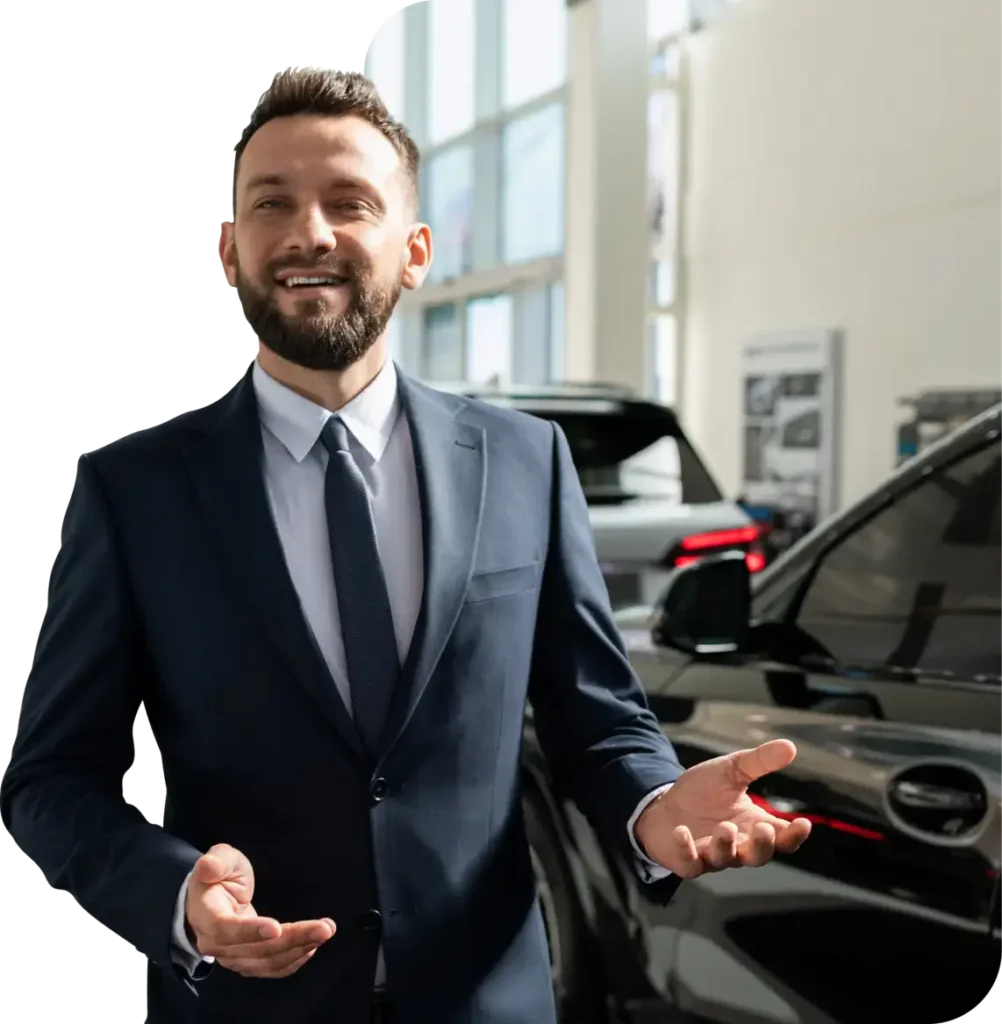 Smiling car salesman in a suit standing inside a dealership next to new vehicles