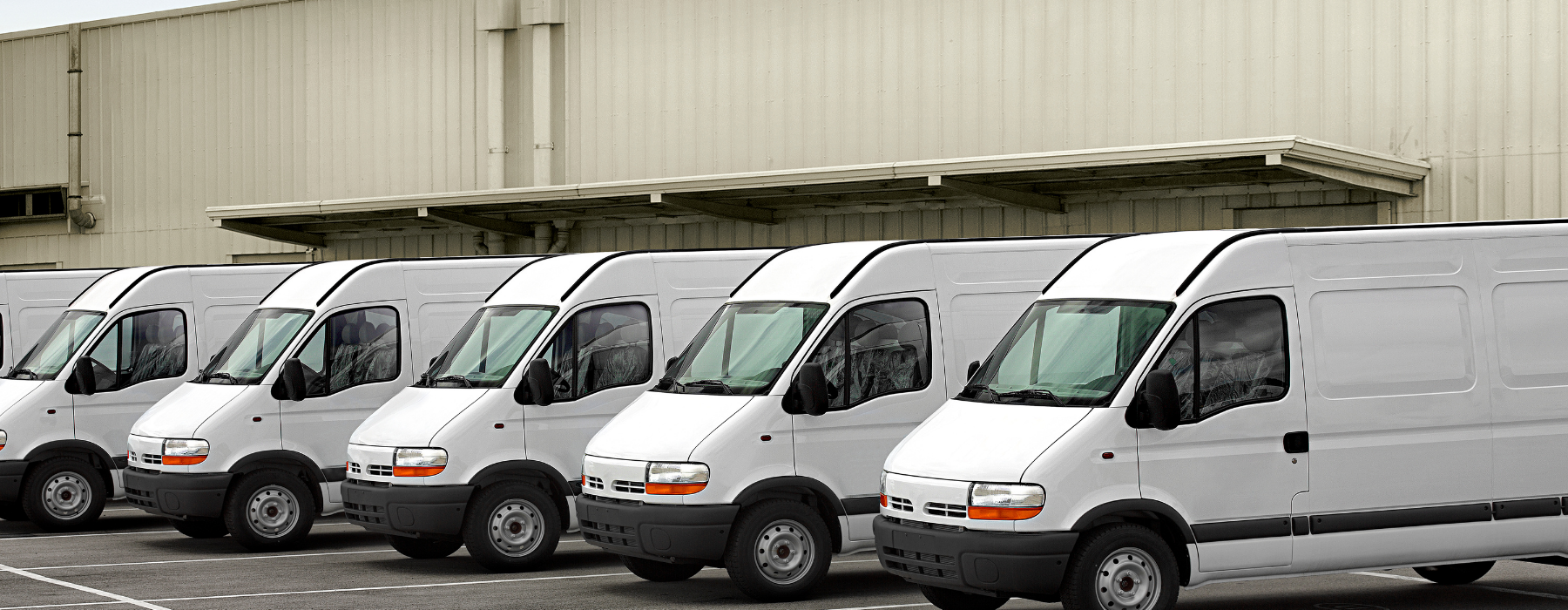 Line of white minibuses parked outside a warehouse, ideal for operators looking for Mini Bus Fleet Insurance to protect multiple vehicles.