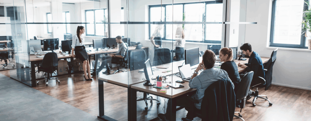 Employees working in a modern open-plan office with glass walls, illustrating the need for Commercial Property Insurance to protect office spaces and business assets.