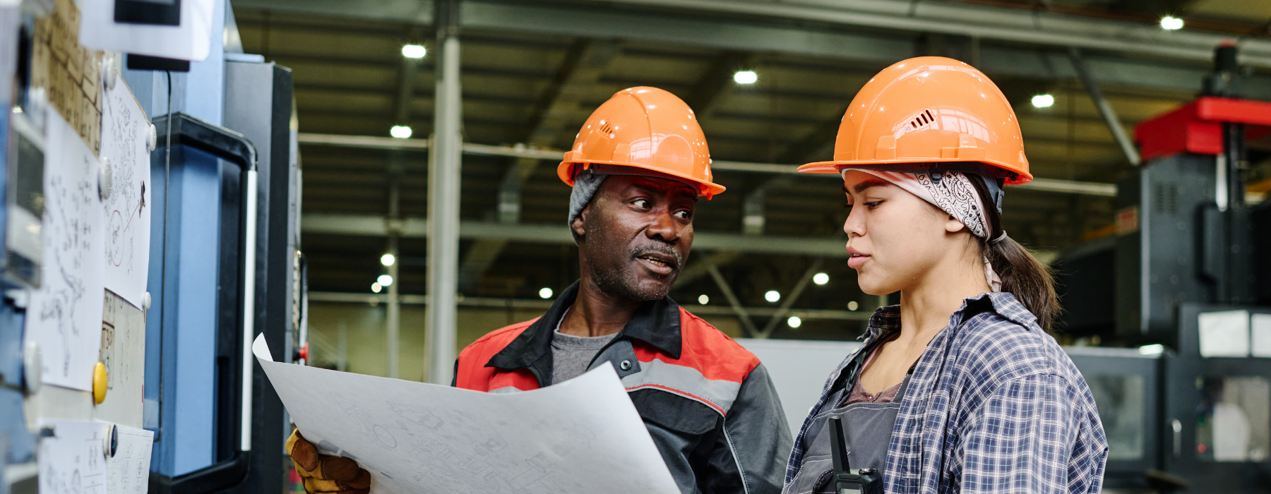 Two factory workers wearing safety helmets reviewing technical plans on a production site, showing the importance of Manufacturer Insurance for staff and operations.