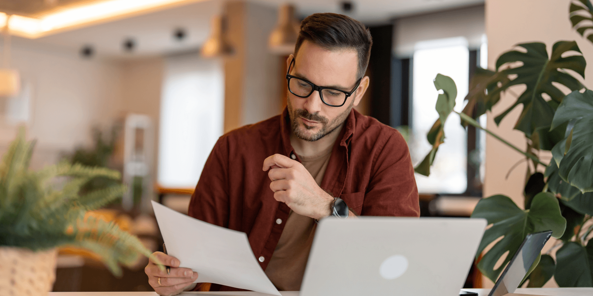 Focused man wearing glasses reviewing paperwork while working on a laptop in a home office.