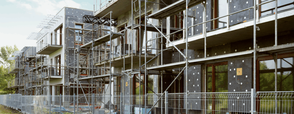 A property development site with new apartment buildings under construction, scaffolding in place, and safety fencing around the area.
