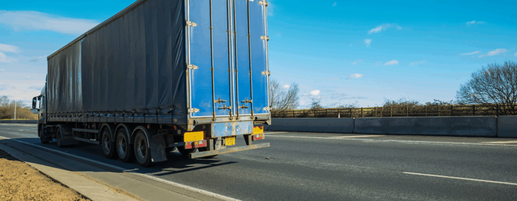 A large blue curtain-sided HGV truck driving on a UK motorway under a clear blue sky, showing the rear view of the vehicle with its trailer and multiple axles.