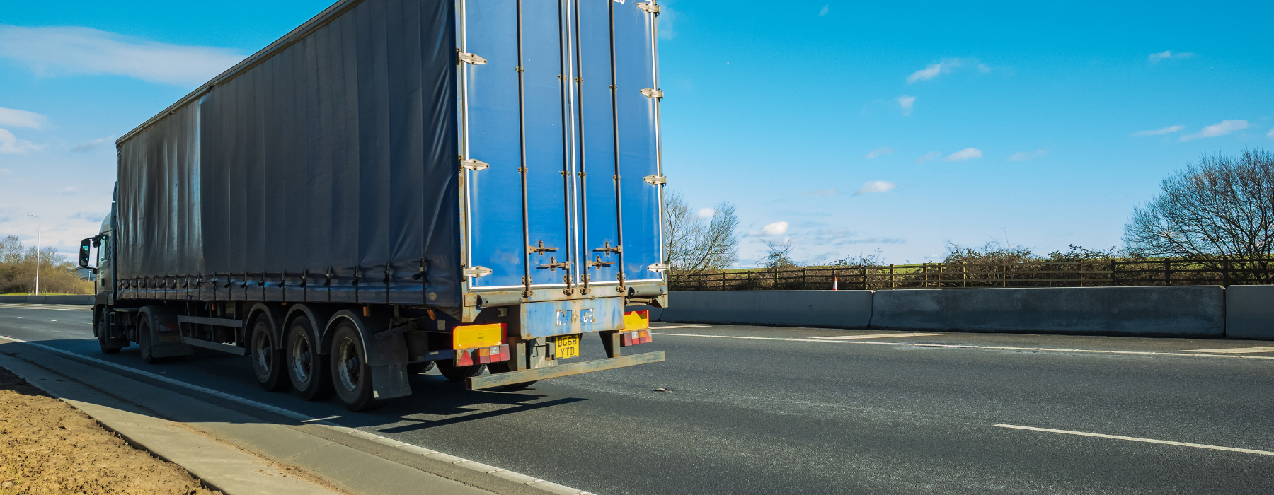A large blue curtain-sided HGV truck driving on a UK motorway under a clear blue sky, showing the rear view of the vehicle with its trailer and multiple axles.