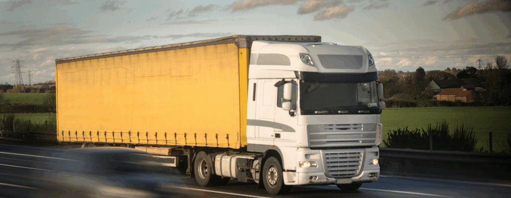 White articulated lorry with a yellow trailer driving on a motorway through the countryside under a cloudy sky.