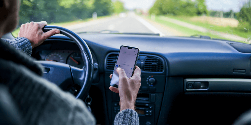 Driver using a mobile phone while driving on a rural road, illustrating distracted driving risk.