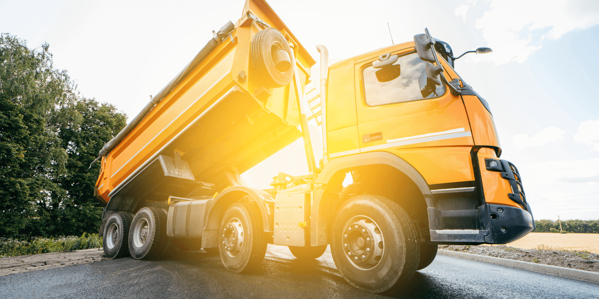 Yellow dumper truck tipping asphalt on a rural road during bright daylight, used for construction and groundwork transport.